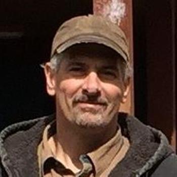 A man in a brown jacket and hat standing outside on a sunny day.