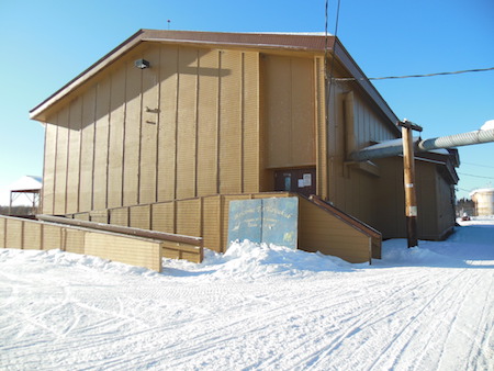 The image  shows a building in the middle of a snowy field. The building is a single story building with a red metal roof. It has a wooden front door and a small window on the right side of the door. There is a ramp leading up to the front door.