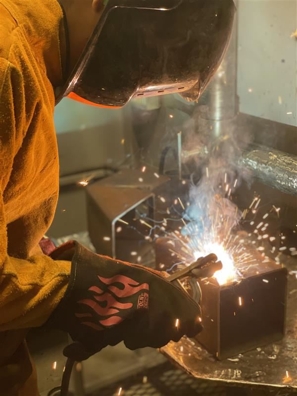 A person in a yellow protective suit with a black welding glove is holding a welding torch close to a metal object, creating bright sparks