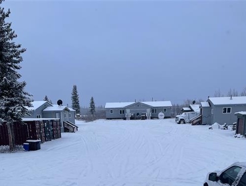 The image shows a white truck parked in a snowy parking lot. The truck appears to be a Ford F-150, a popular pickup truck model. It is white and has a long bed. The truck is parked in a large parking lot that is covered in snow. There are several other vehicles parked in the lot, but they are all far away from the white truck. In the background, there are trees and a building.