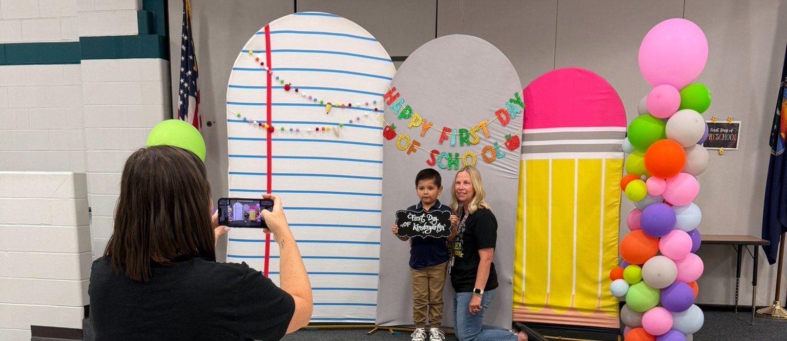 A woman takes a photo of a young child in front of a colorful backdrop with a pencil-shaped balloon.