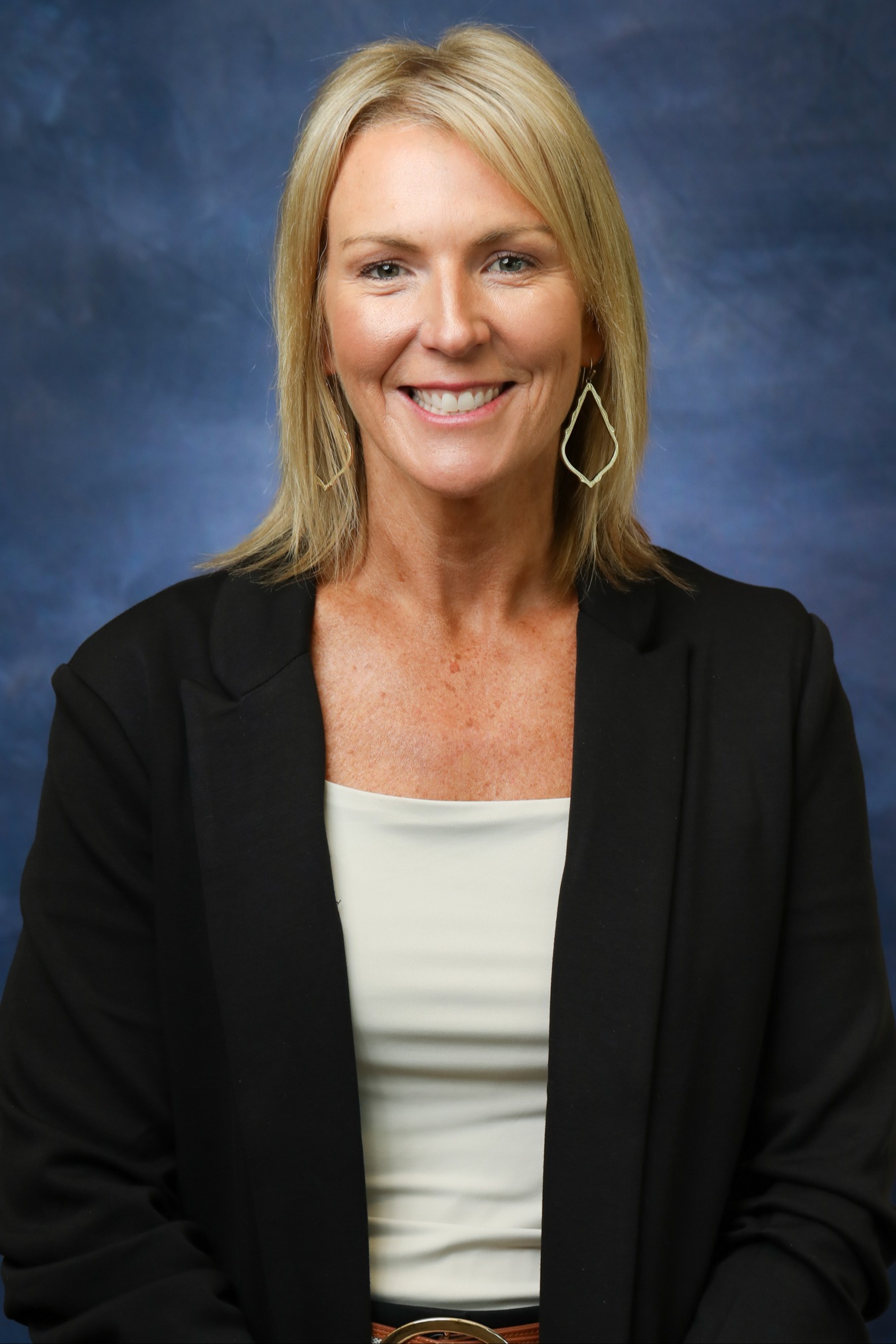A woman with blonde hair and teardrop outline earrings wearing a black blazer smiling