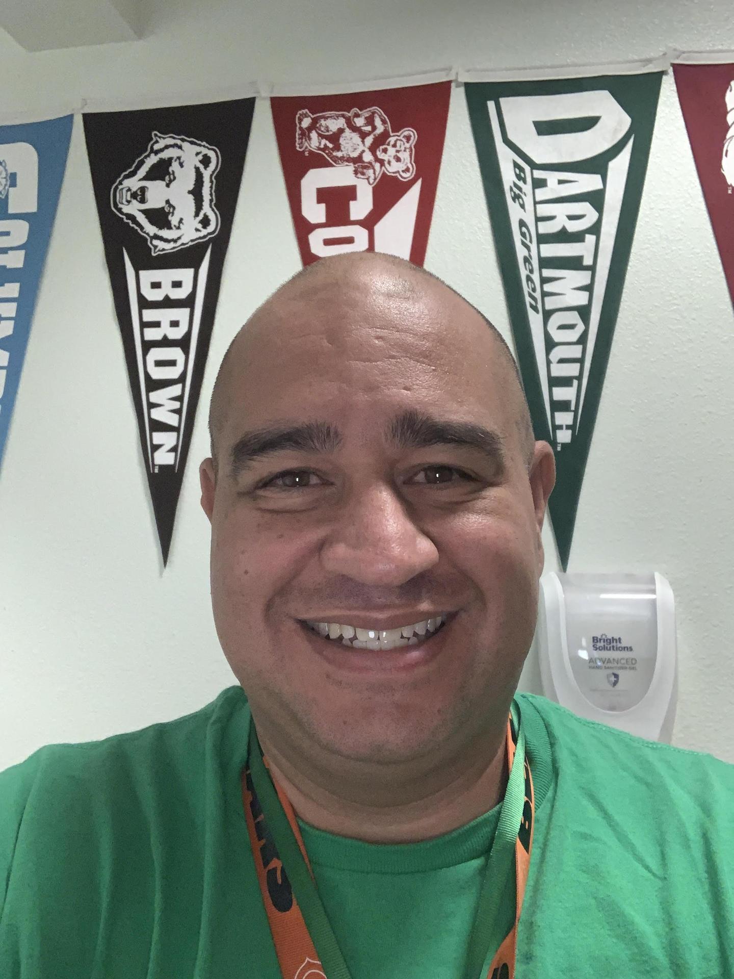 A man with a shaved head smiling in front of college pennants hanging on a wall.
