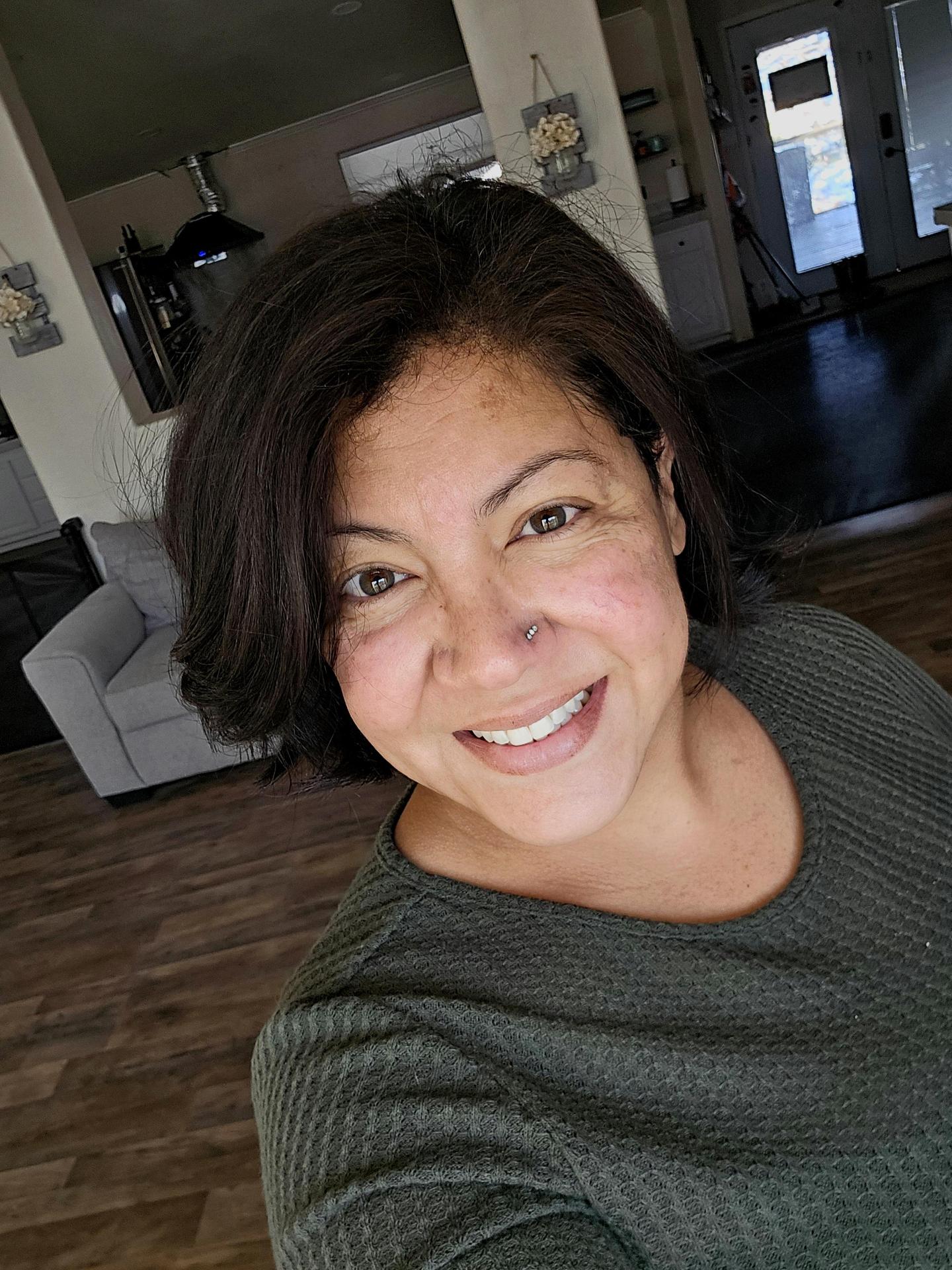 A woman with dark hair and a nose piercing smiling in a home interior.
