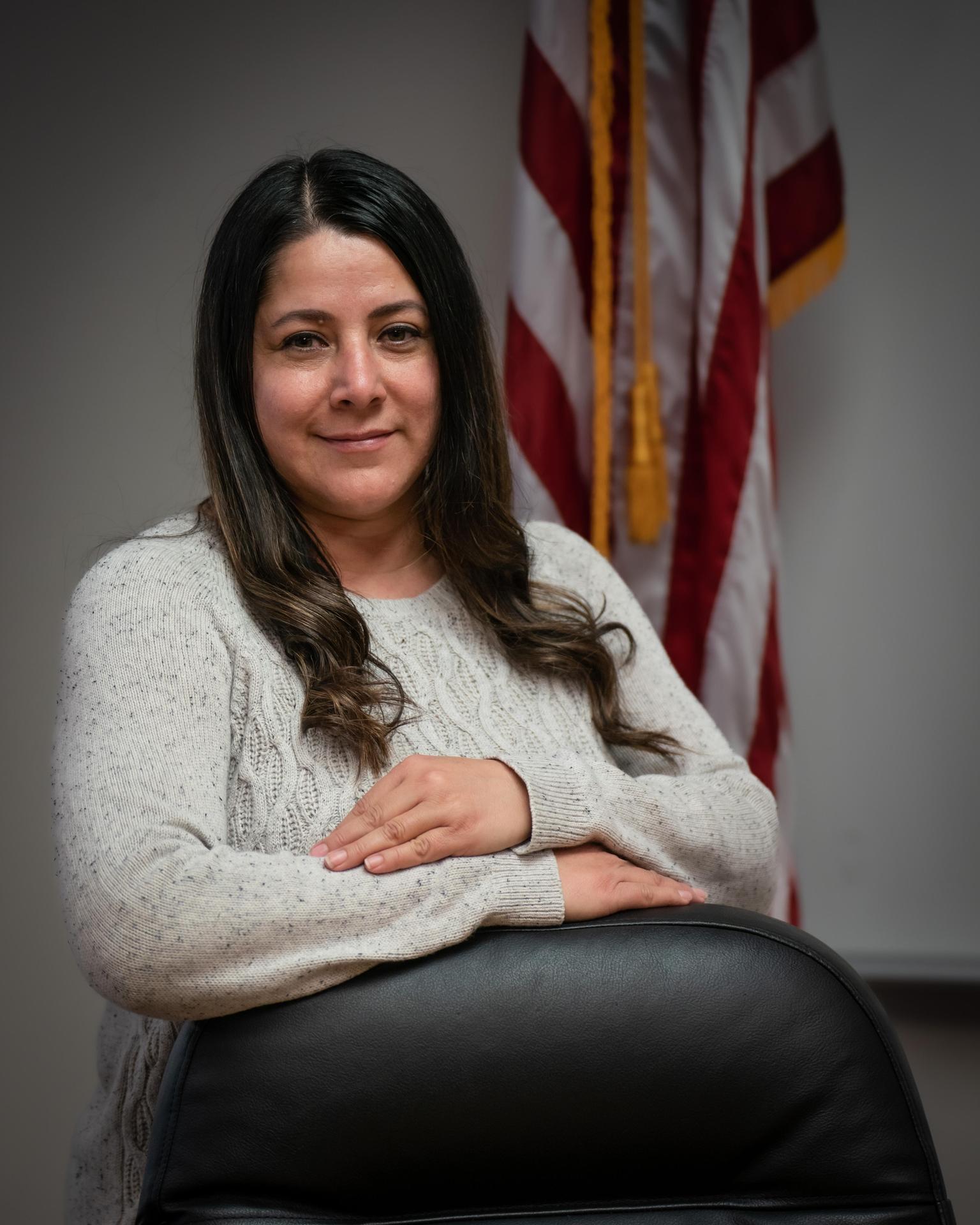 A woman with long dark hair wearing a light-colored sweater, sitting with her arms crossed in front of an American flag.