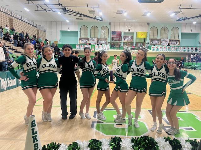 A cheerleading team in green and white uniforms stands on a basketball court with a coach, posing for a photo.