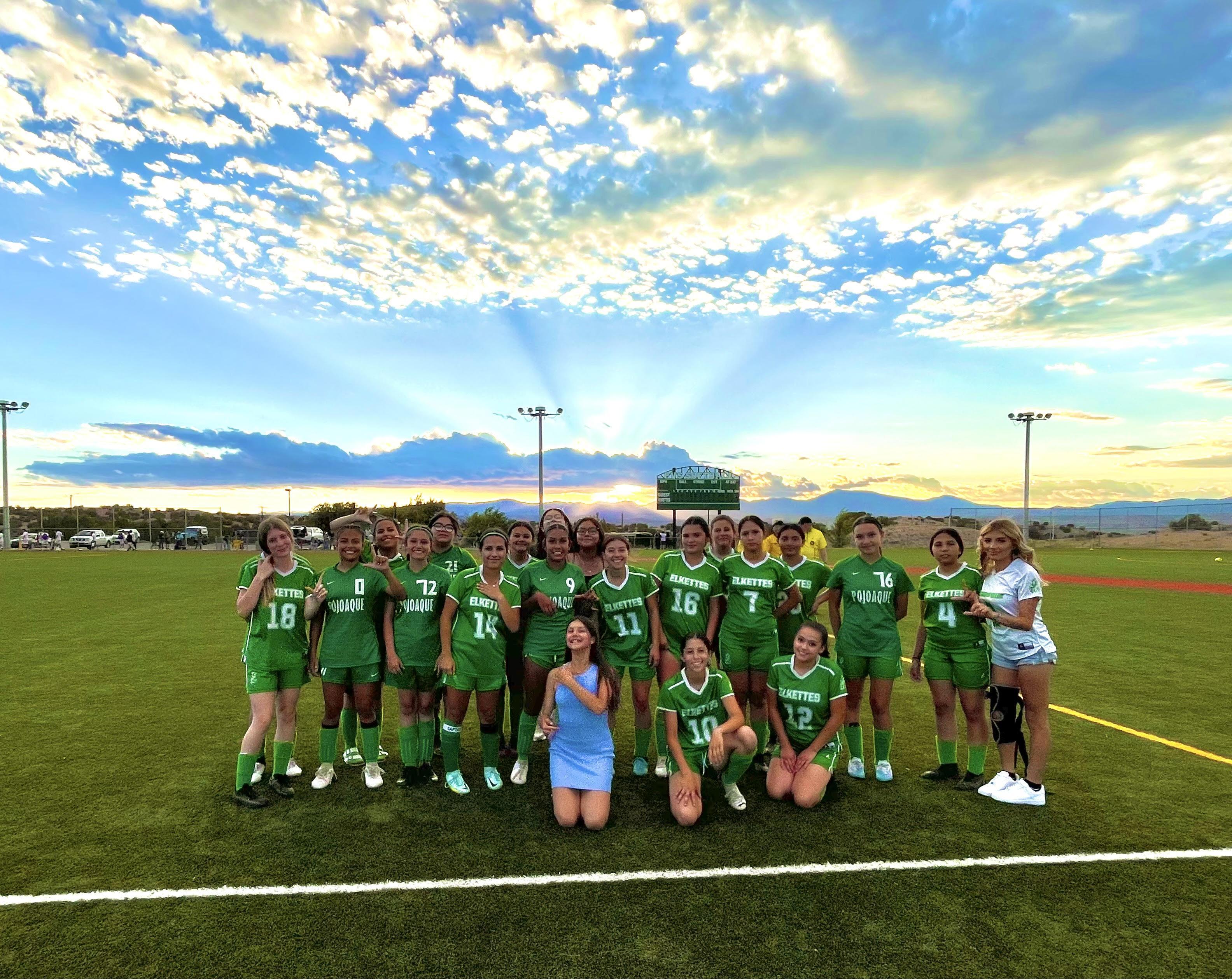 A girls' soccer team in green uniforms poses on a field at sunset with a coach, some kneeling and others standing.