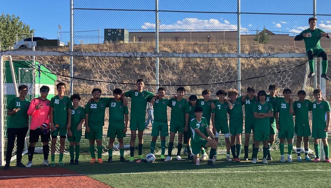 A soccer team in green uniforms stands in a line on a field with a goal behind them, with a coach and a goalkeeper on the sides.