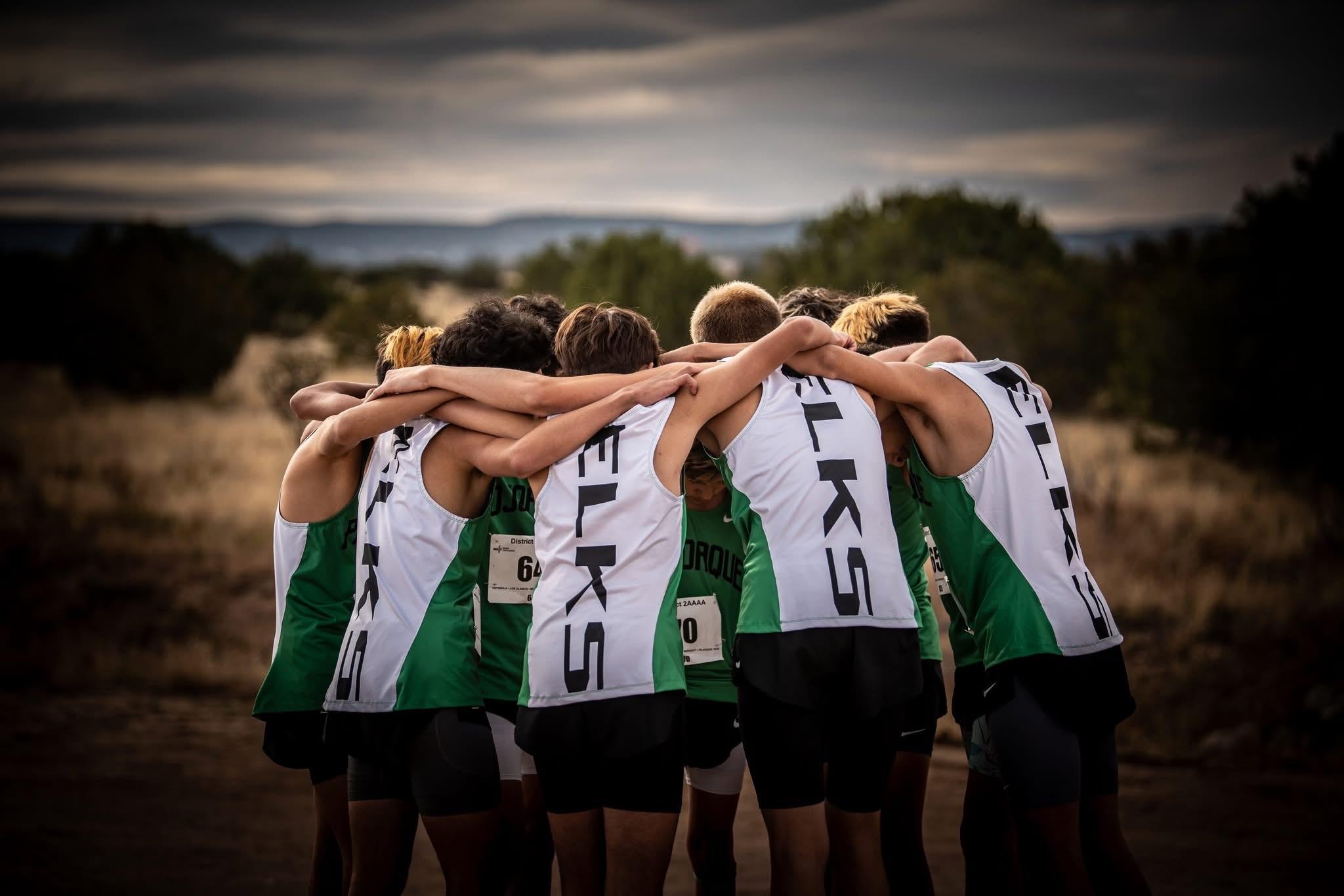 A group of athletes in green and white uniforms huddle together with arms around each other outdoors.