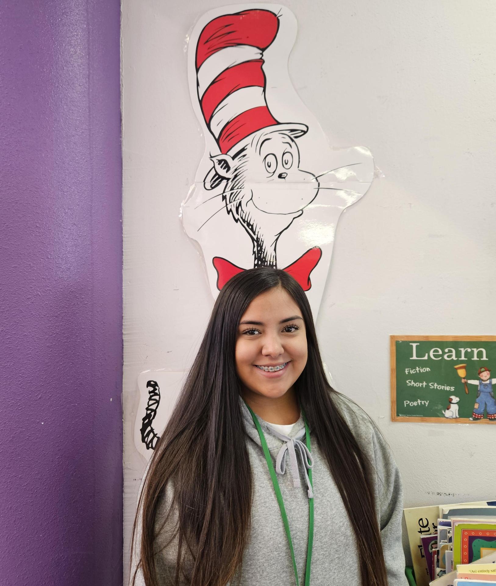 A young woman with long dark hair smiling in front of a wall with a large Dr. Seuss character, the Cat in the Hat, above her.