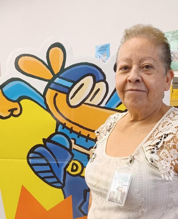 An elderly woman with short hair and a lace top stands next to a colorful cartoon character of a bee dressed as a superhero.