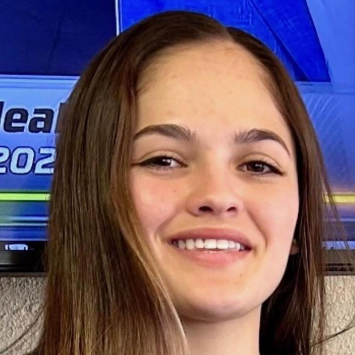 A young woman with long brown hair smiling at the camera, wearing a black shirt and a necklace with a circular pendant.