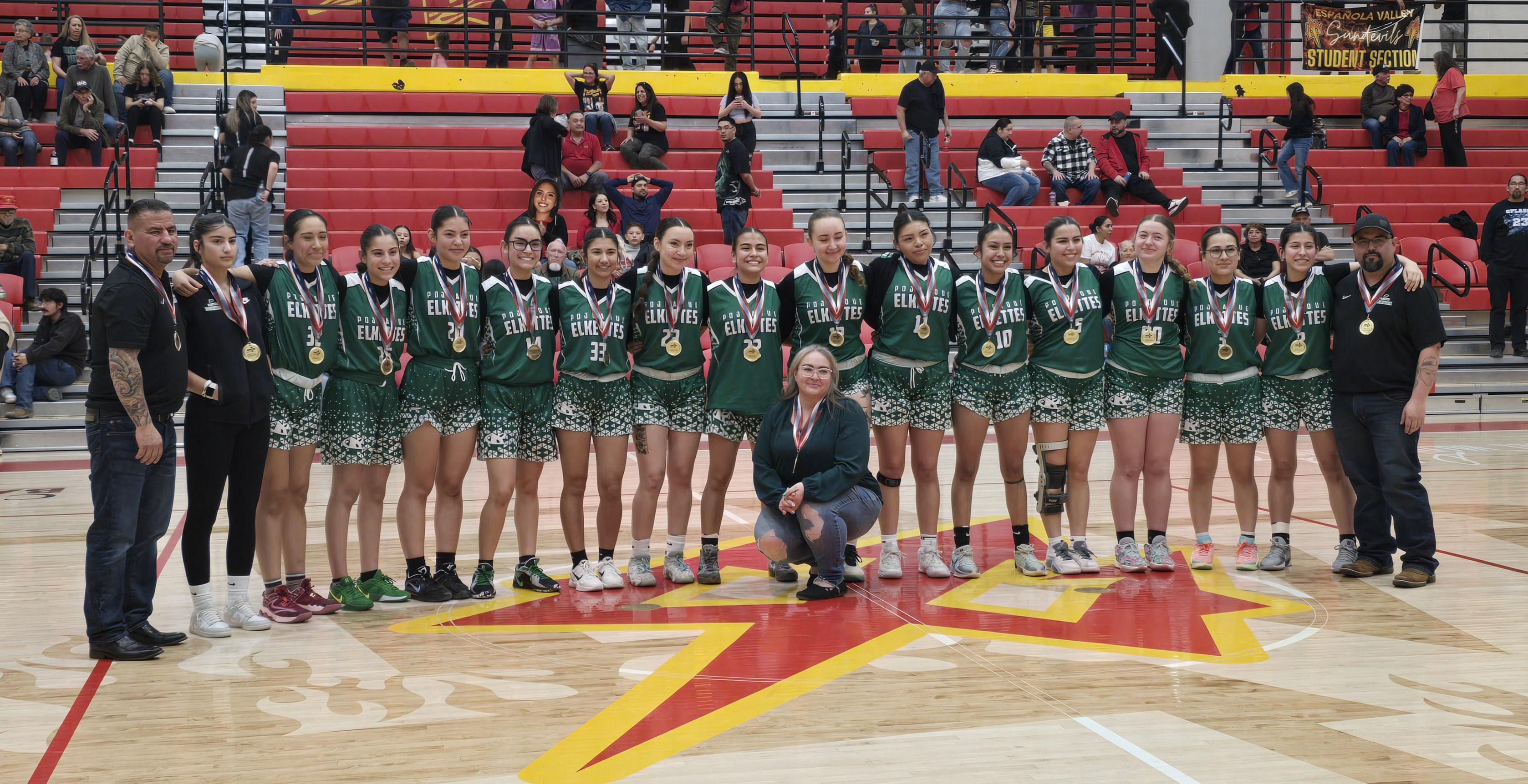 A girls' basketball team in green uniforms stands with coaches on a basketball court, posing for a team photo.