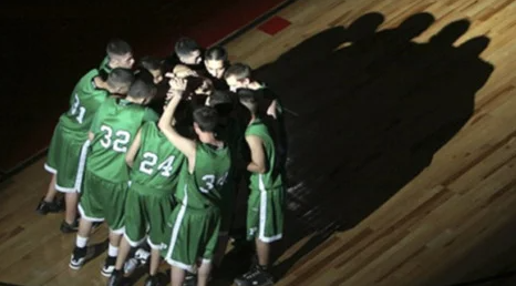 A basketball team in green uniforms huddles together on the court, with their shadows cast on the wooden floor.