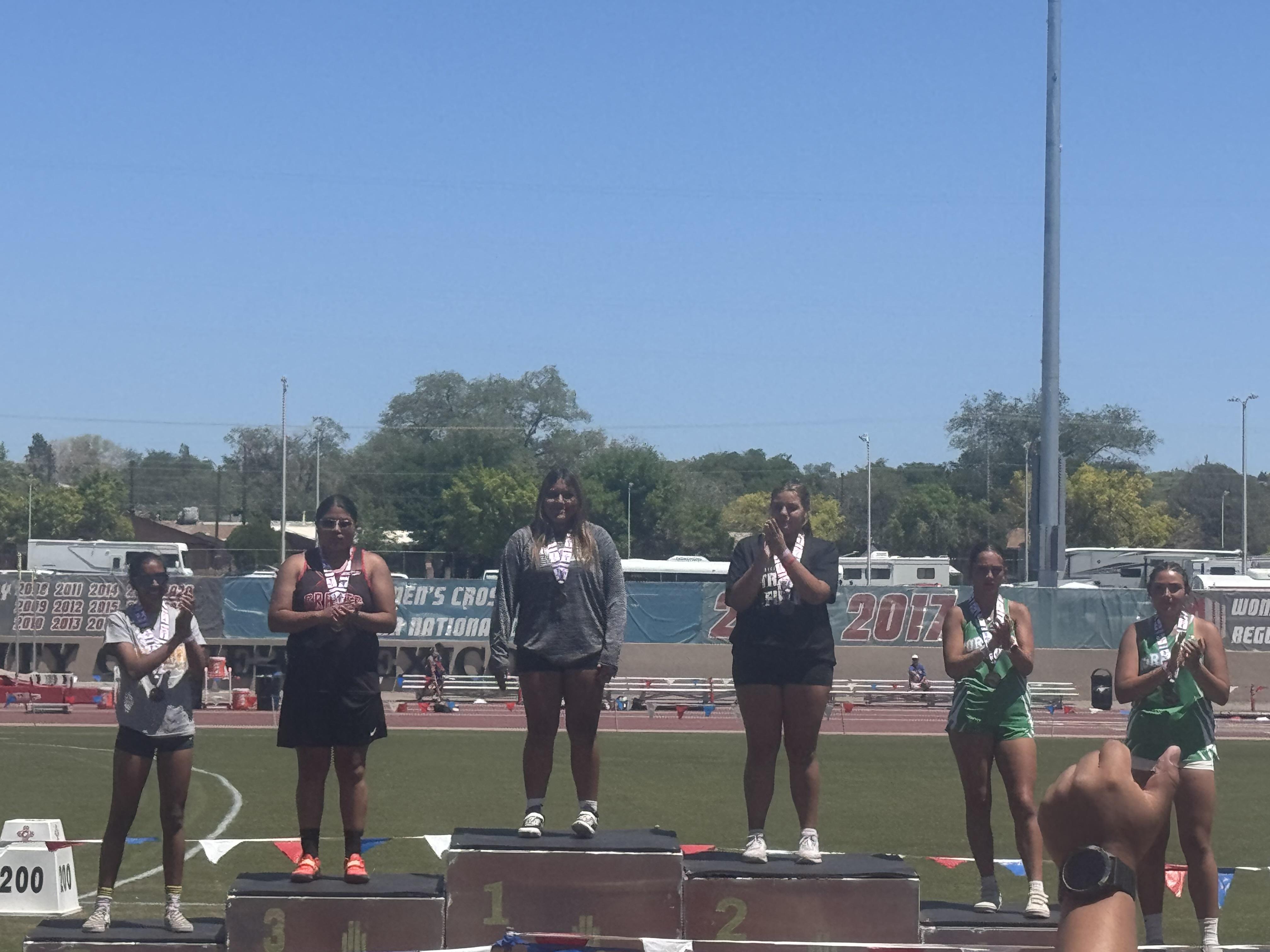Six female athletes stand on a winners' podium at an outdoor track and field event, with some holding medals.