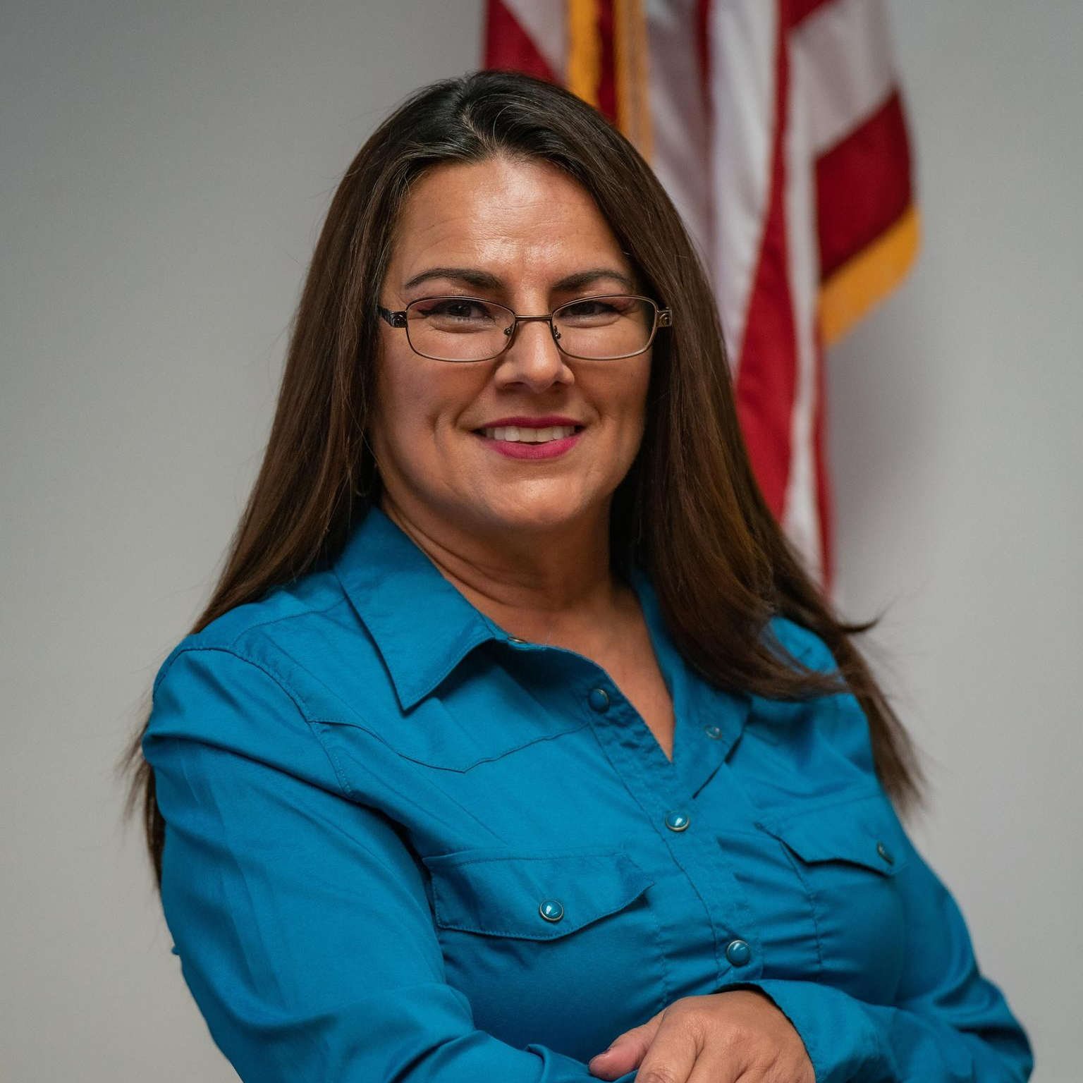 A woman with long brown hair, glasses, and a blue shirt stands in front of an American flag, smiling with arms crossed.