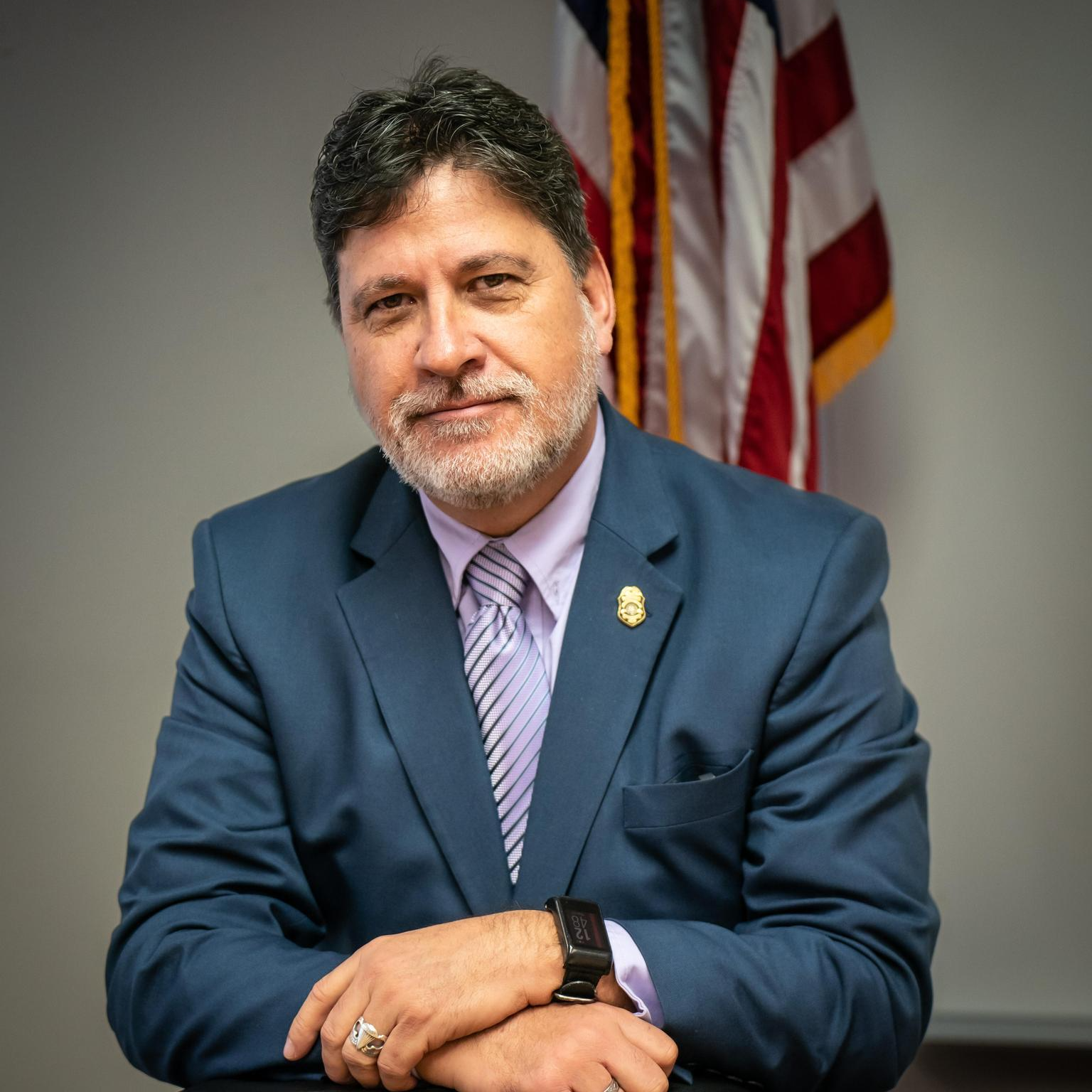 A man with gray hair and a beard, wearing a blue suit and purple tie, sits with his arms crossed in front of an American flag.