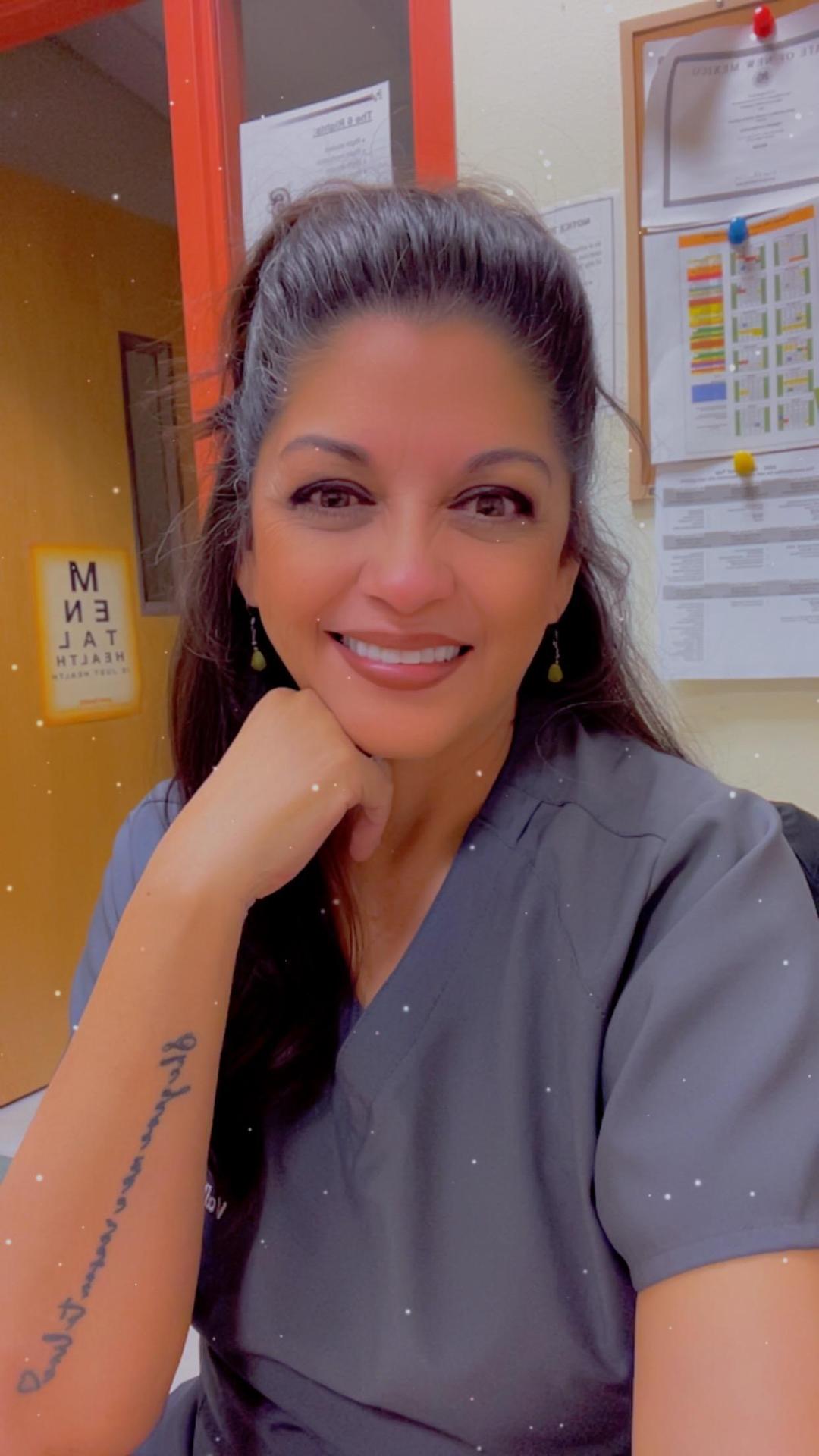 A woman with long dark hair smiling and resting her chin on her hand in an indoor setting.