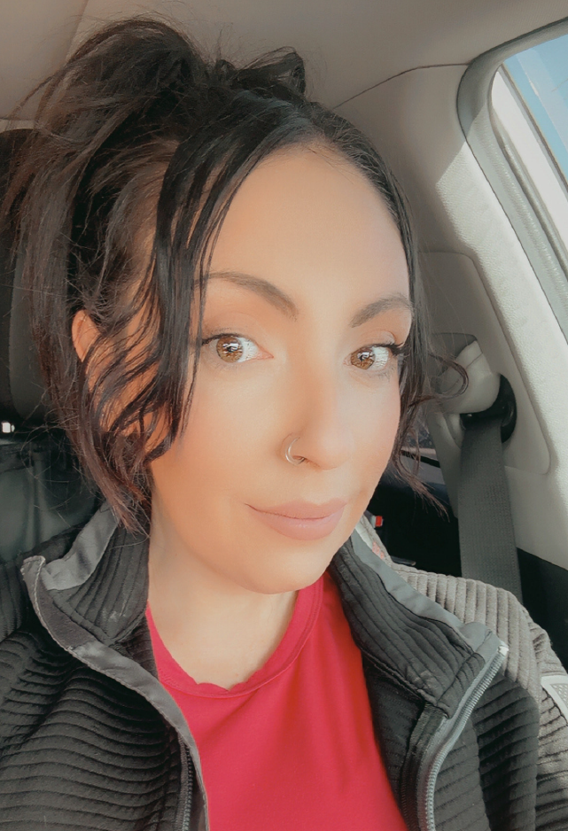 A woman with dark wavy hair, light skin, and a nose piercing, wearing a red shirt and black jacket, sitting in a car.