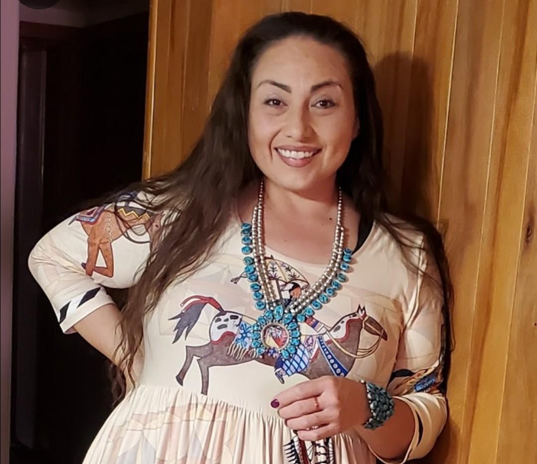 A woman with long dark hair smiling and wearing a colorful dress with horse illustrations, accessorized with necklaces and bracelets, standing against a wooden wall.