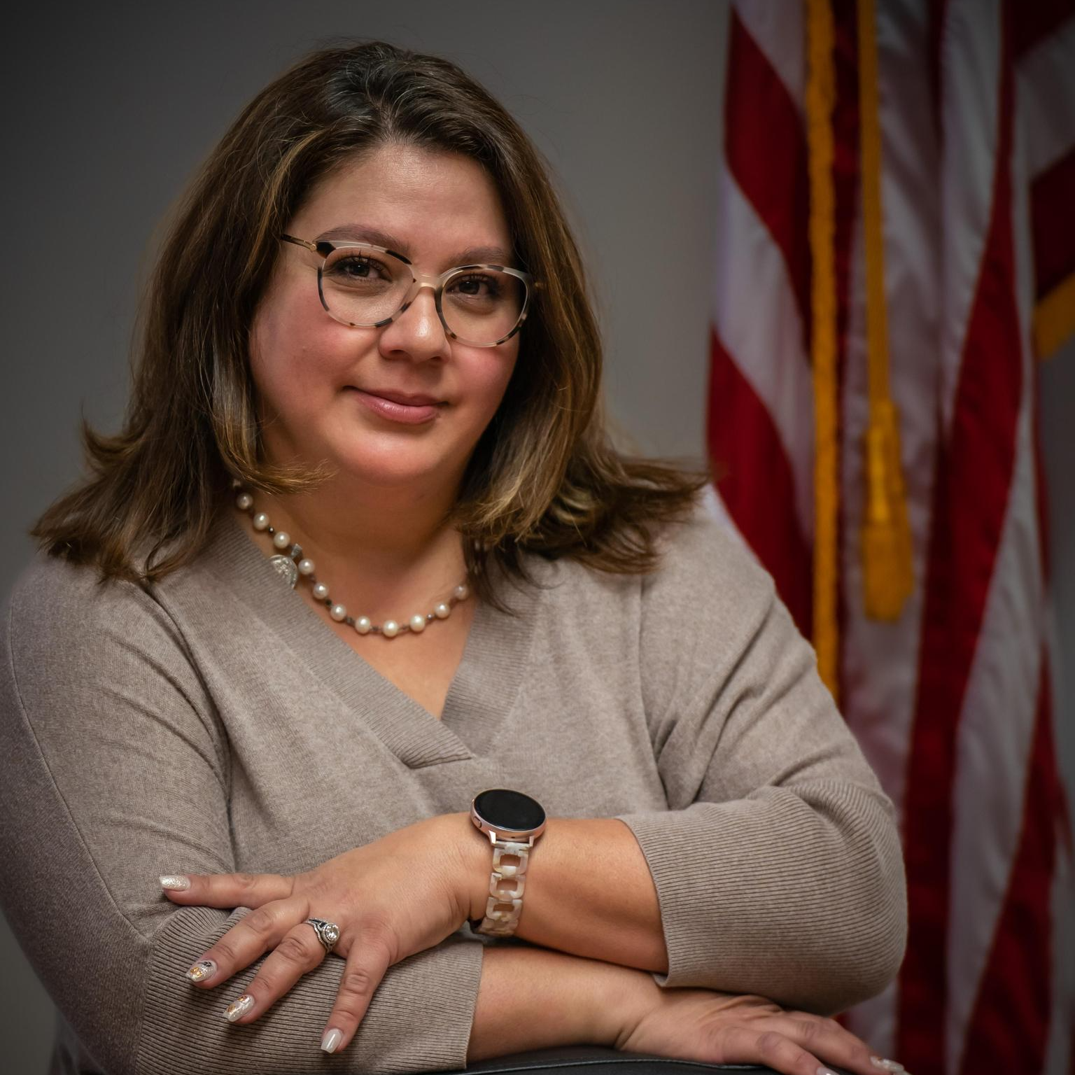 A woman with shoulder-length brown hair, glasses, and a pearl necklace, sitting with arms crossed in front of an American flag.