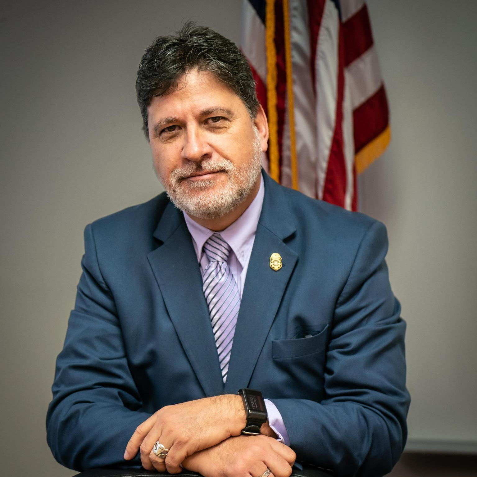 A man in a blue suit with a purple tie, sitting with arms crossed, in front of an American flag.