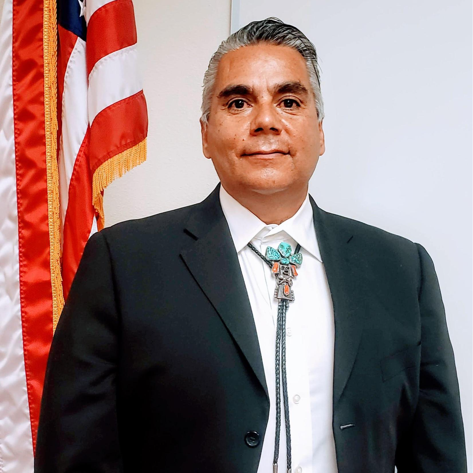 A man in a black suit and white shirt stands in front of an American flag, wearing a turquoise bolo tie, with a neutral expression.