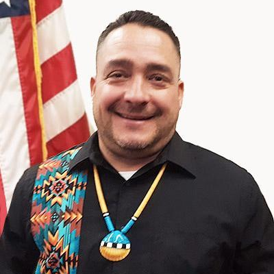 A man smiling in front of an American flag, wearing a black shirt with a colorful patterned section and a beaded necklace with a pendant.