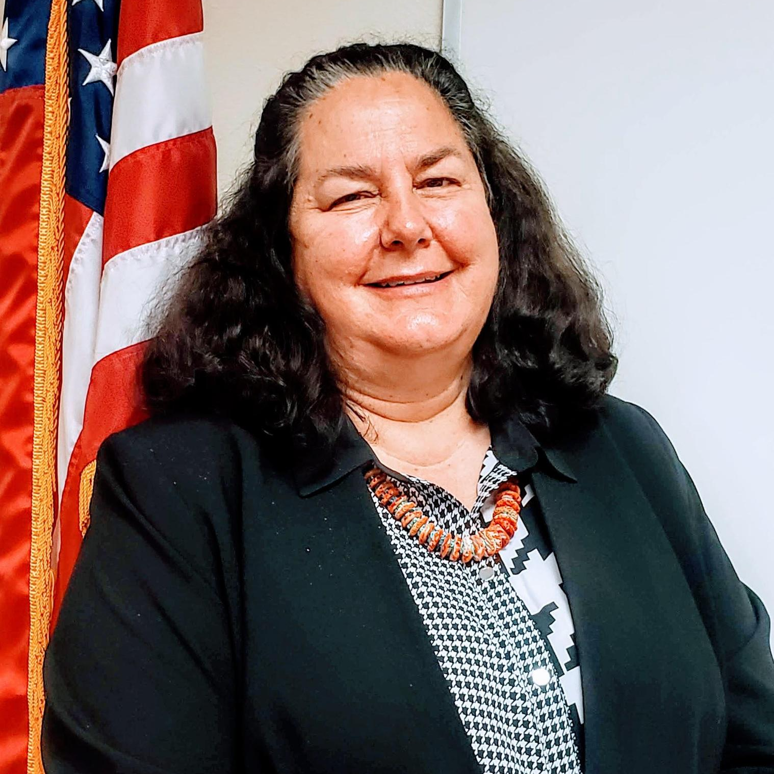 A woman with dark, curly hair, smiling and winking, standing in front of an American flag.