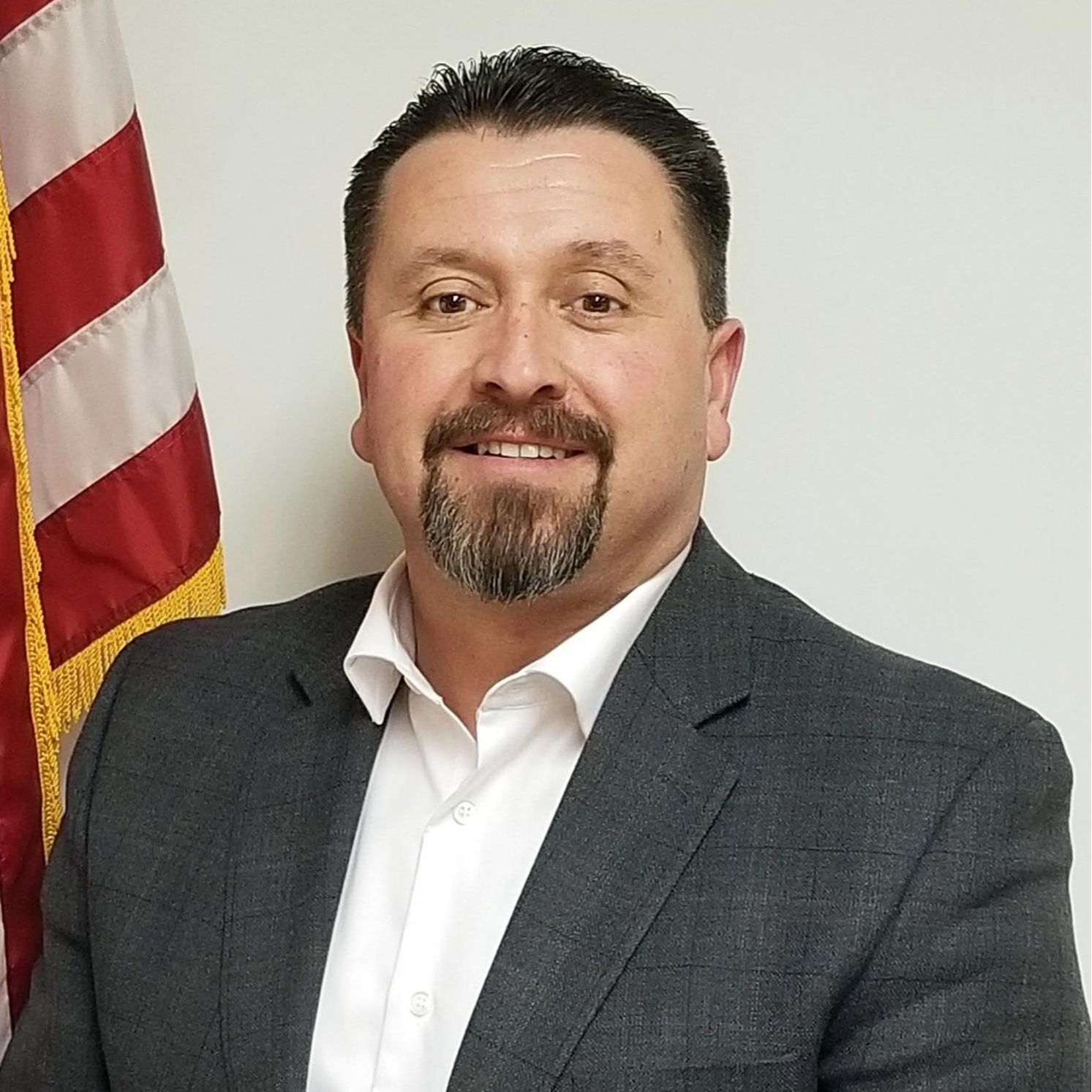 A man with dark hair and a beard, wearing a gray suit and white shirt, standing in front of an American flag.