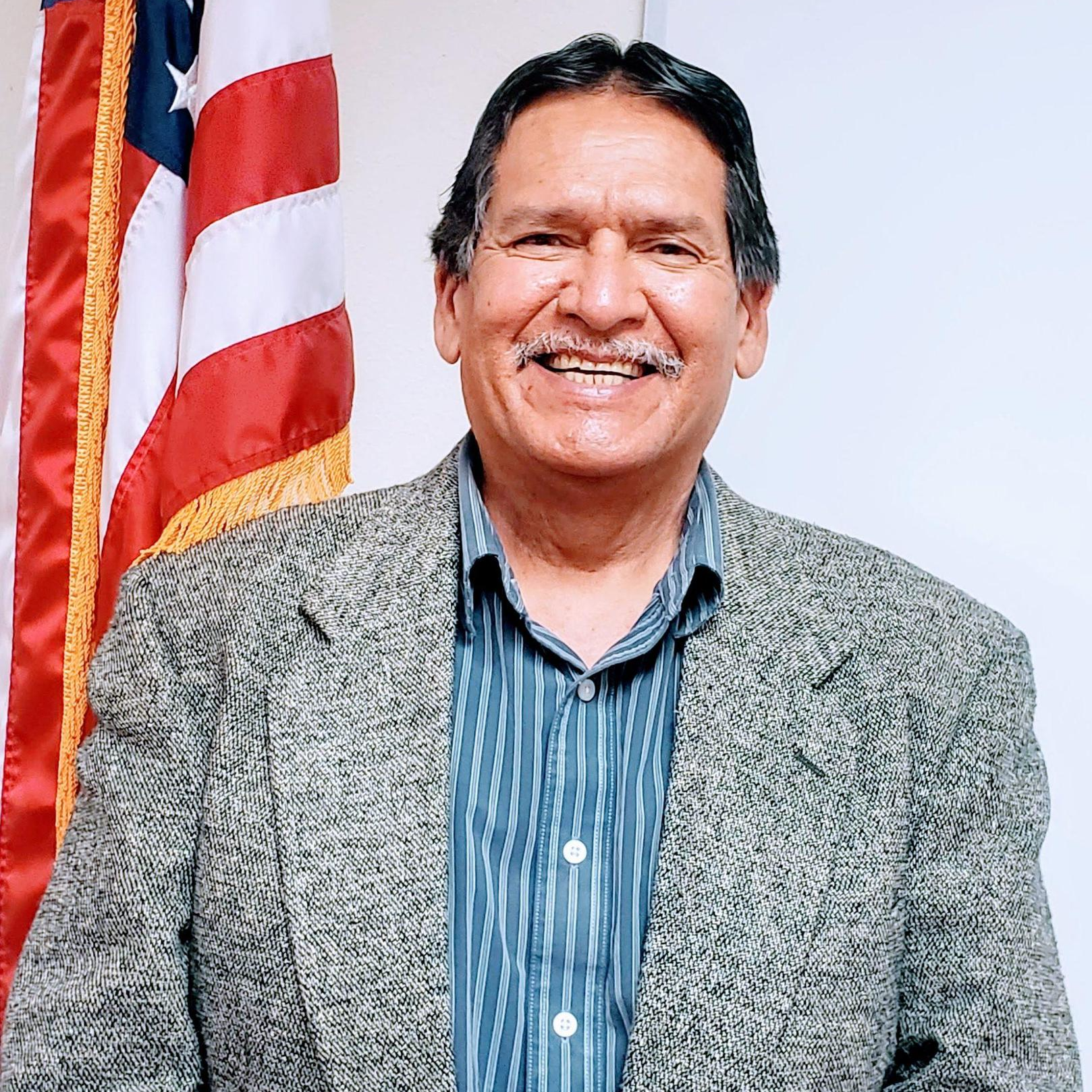 A smiling man in a gray blazer and blue striped shirt stands in front of an American flag.