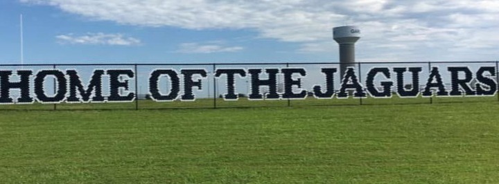 A sign reads "Home of the Jaguars" over a wide, green field, with a water tower in the background.