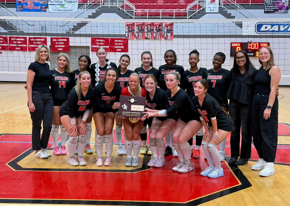 Volleyball team wearing black uniforms stands on a basketball court with a red floor, holding a trophy.
