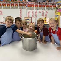 A group of children in aprons gathered around a large pot, engaged in a cooking activity.