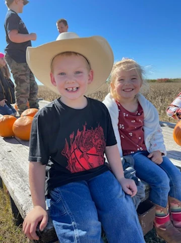 Two kids in cowboy hats sitting on a bench in a pumpkin patch