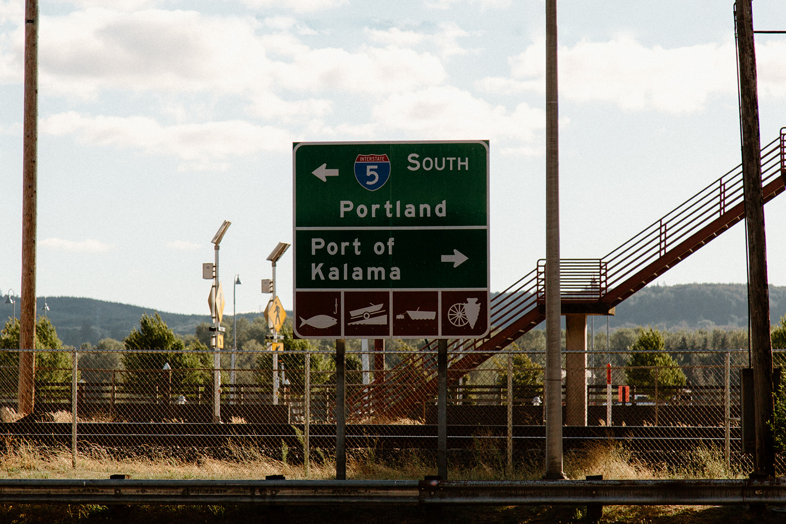 This image shows a green and white metal highway sign with black text. The sign is mounted on a metal post on the side of a highway.