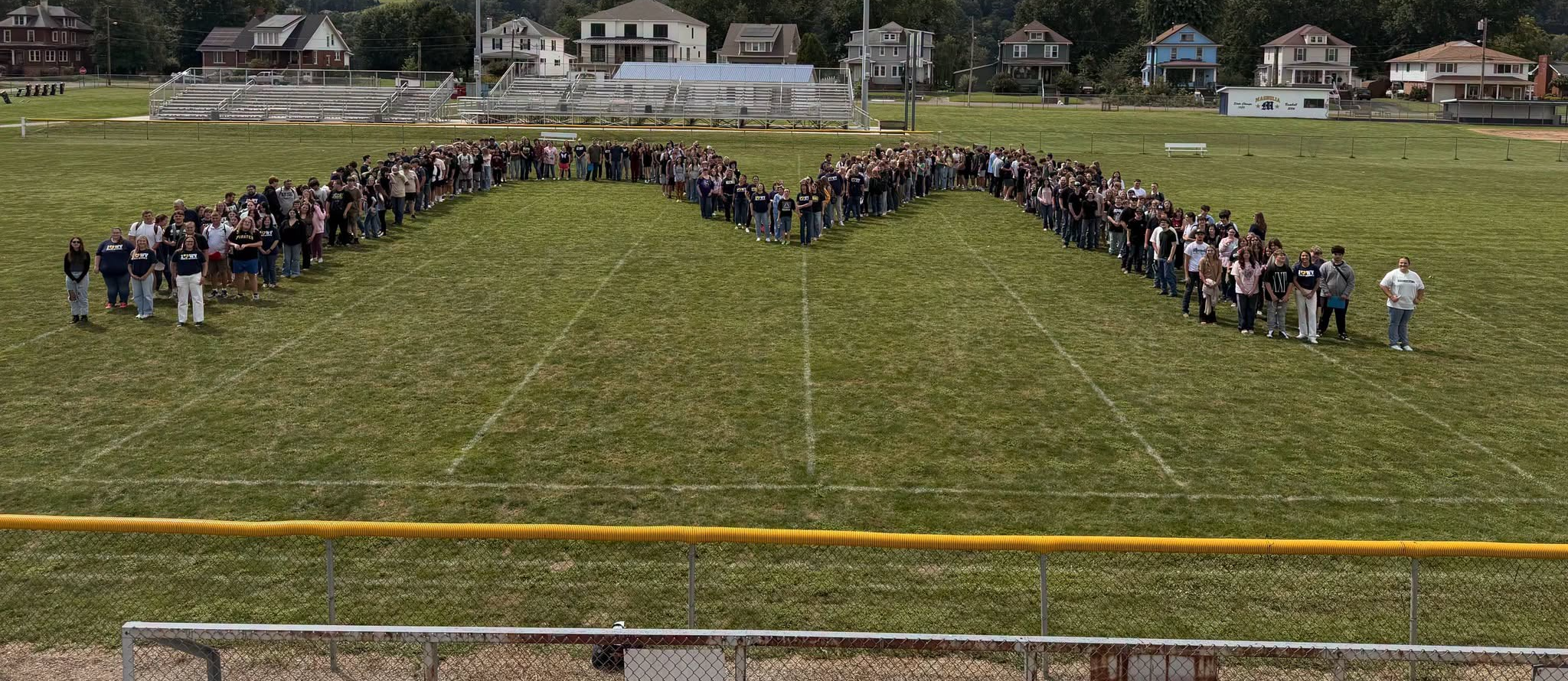 A group of people stand on a football field in a U-shape formation. In the background, houses and mountains are visible under a partly cloudy sky.