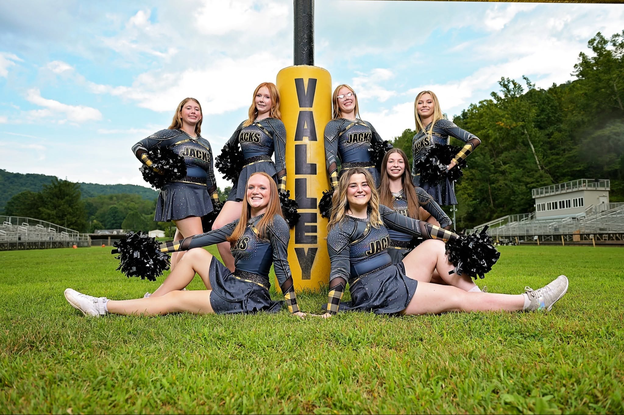 A cheerleading team in uniforms poses on a field, with a yellow "VALLEY" sign behind them and a stadium in the background.
