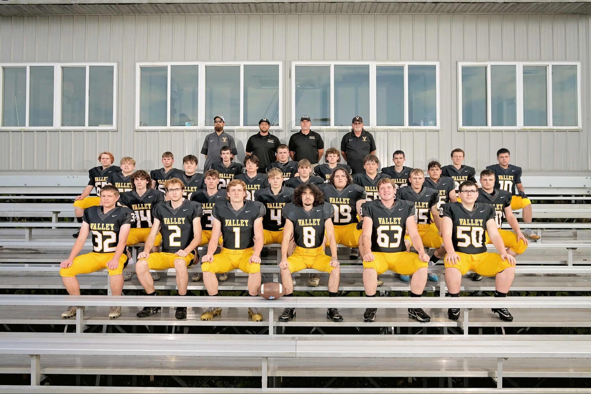 Football team in black jerseys with yellow numbers on bleachers, two coaches standing behind.
