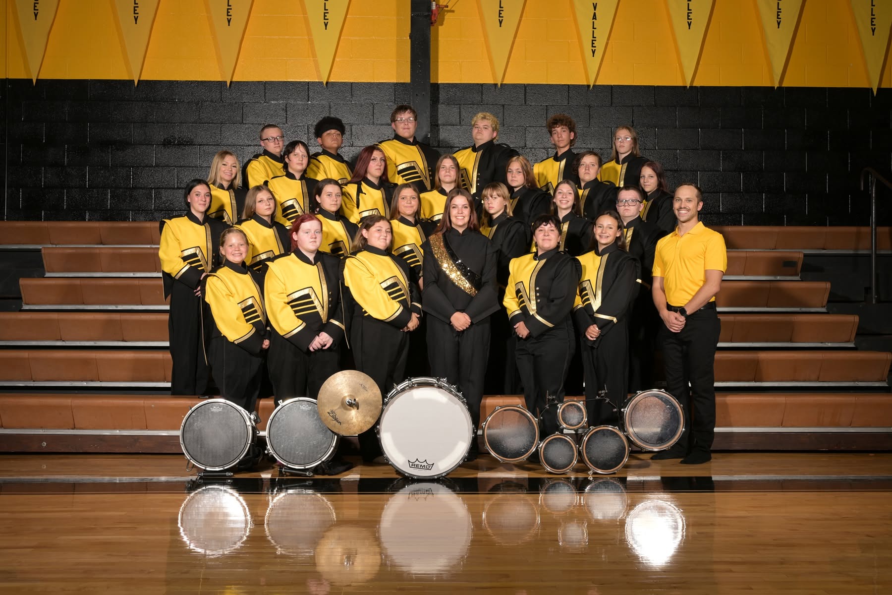 A group of band members, wearing black and yellow uniforms, standing in a gym with instruments on the floor.