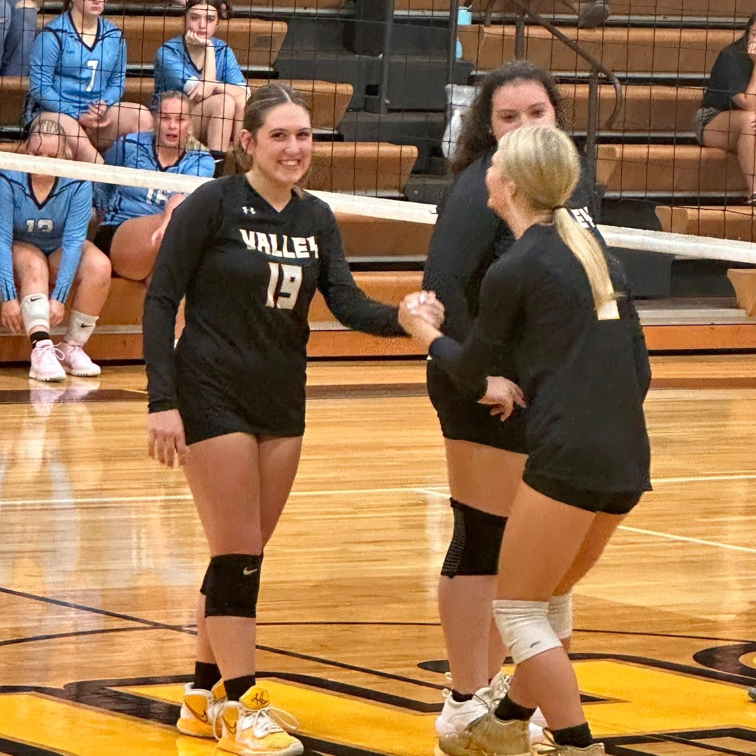 Three female volleyball players in black uniforms shake hands on a court. Spectators are seated on bleachers in the background.