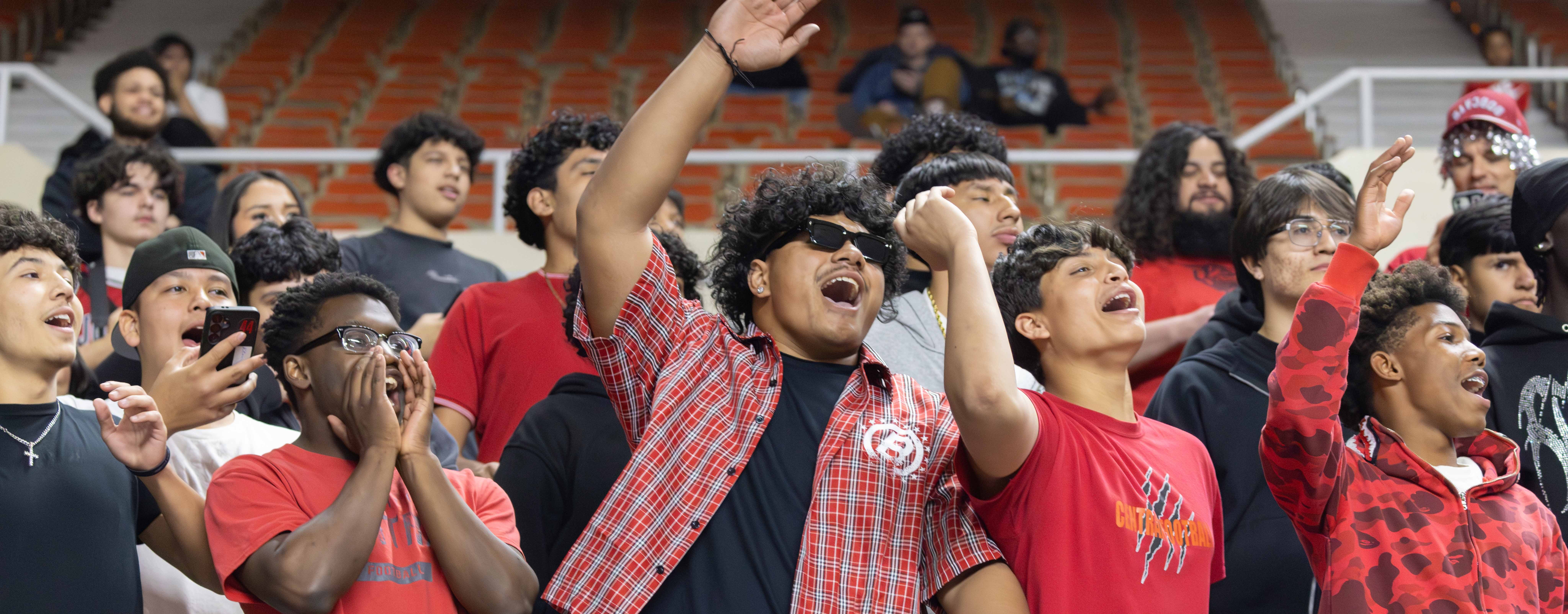 A group of young people in a stadium, wearing red and black shirts, cheering with their hands up.