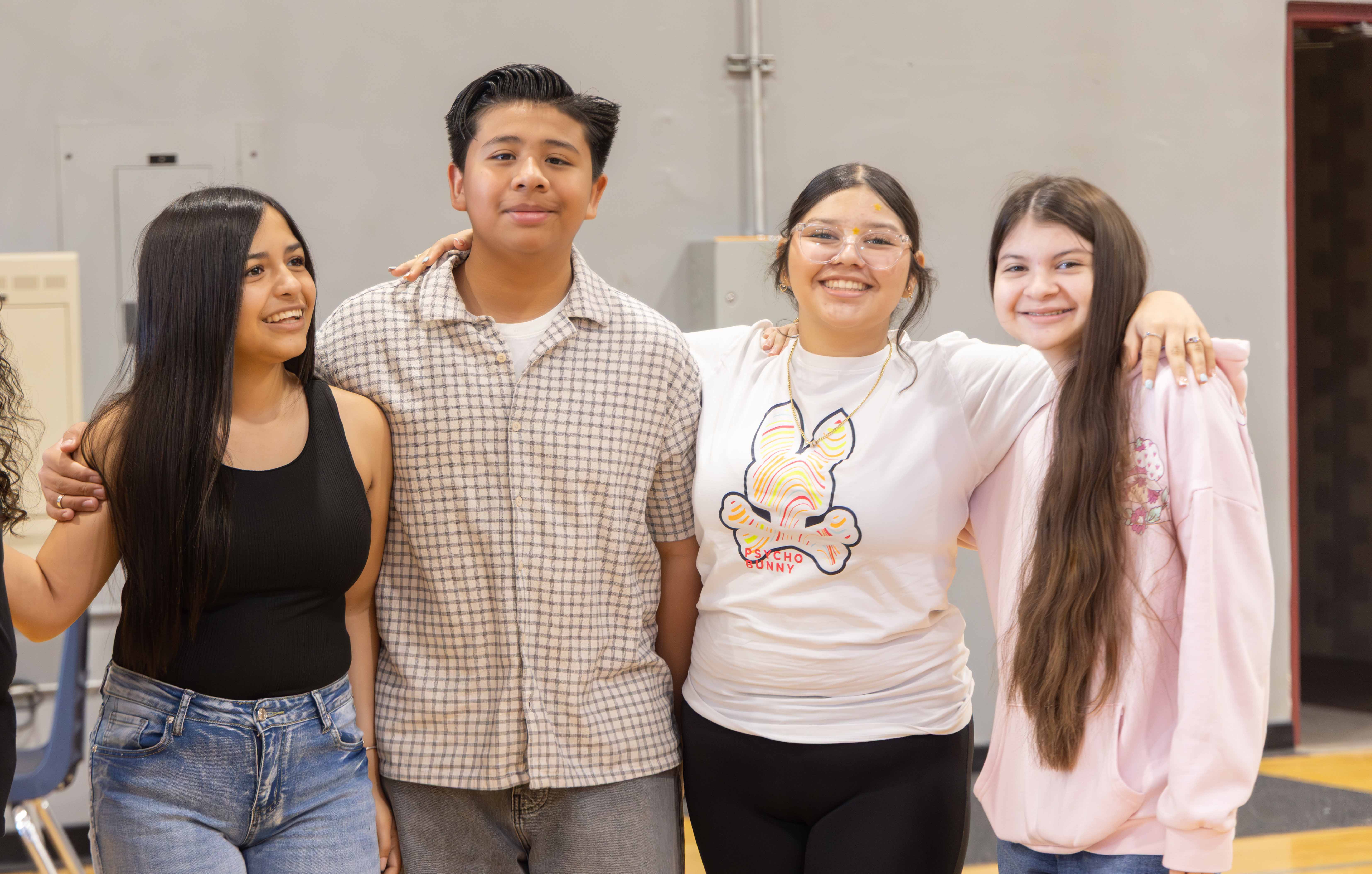 Four young people stand together; two women and two men. They wear casual clothing. Background is a room with light walls and a wooden floor.