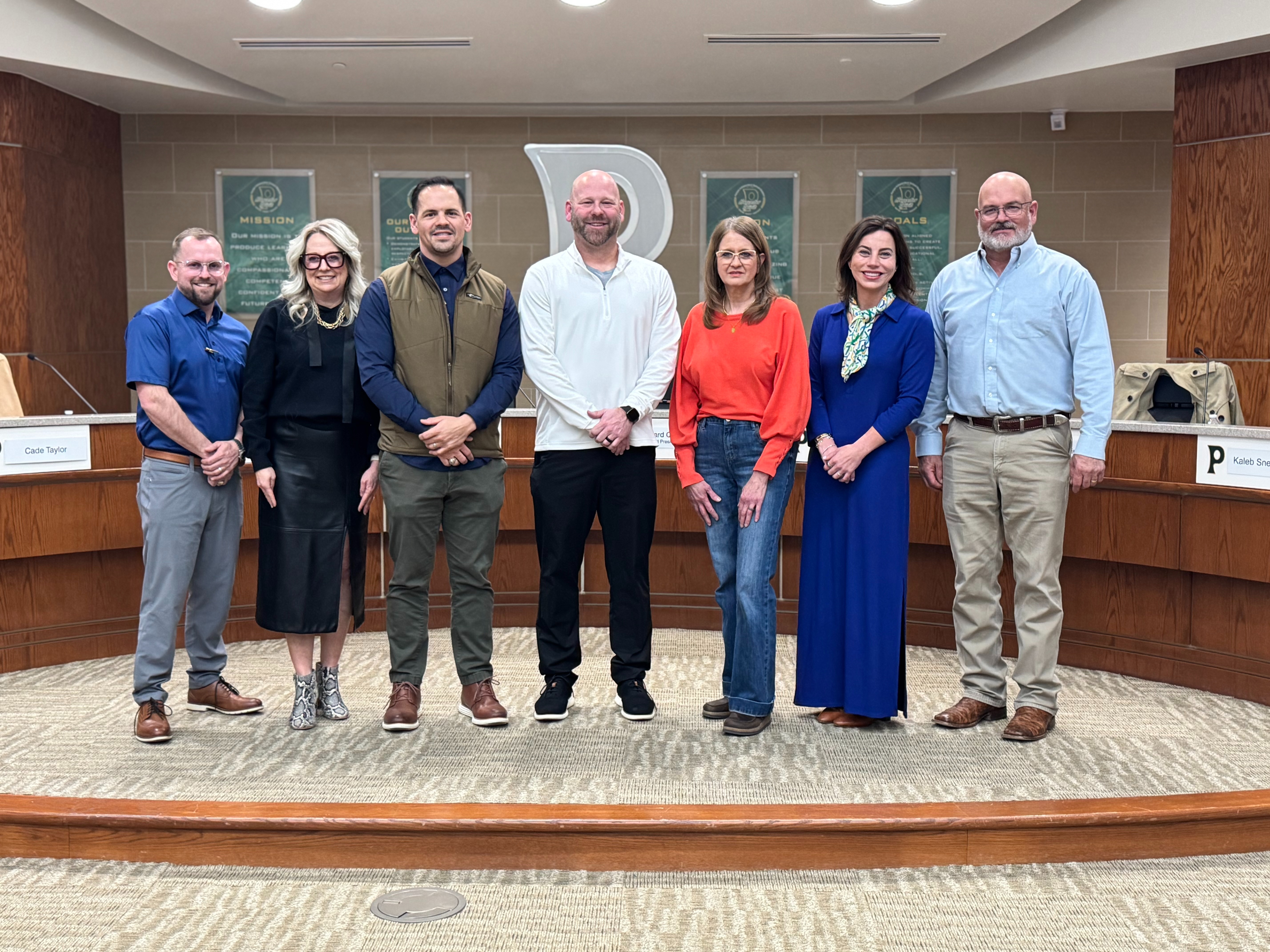 A photo of seven men and women standing in front of a School logo