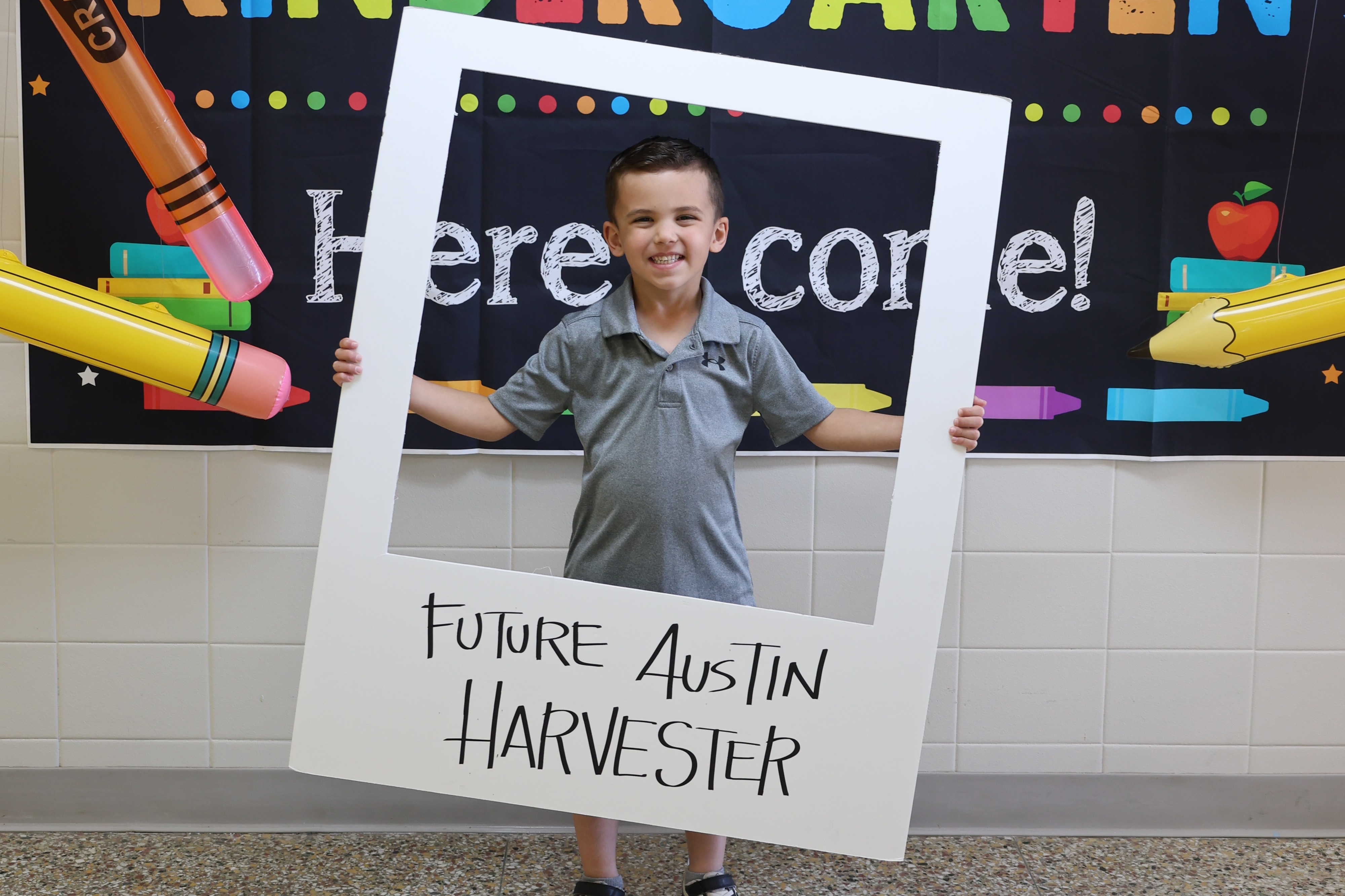 A young boy stands in front of a bulletin board reading "Watch Out Kindergarten" and holds a sign that says "Future Harvester Austin".