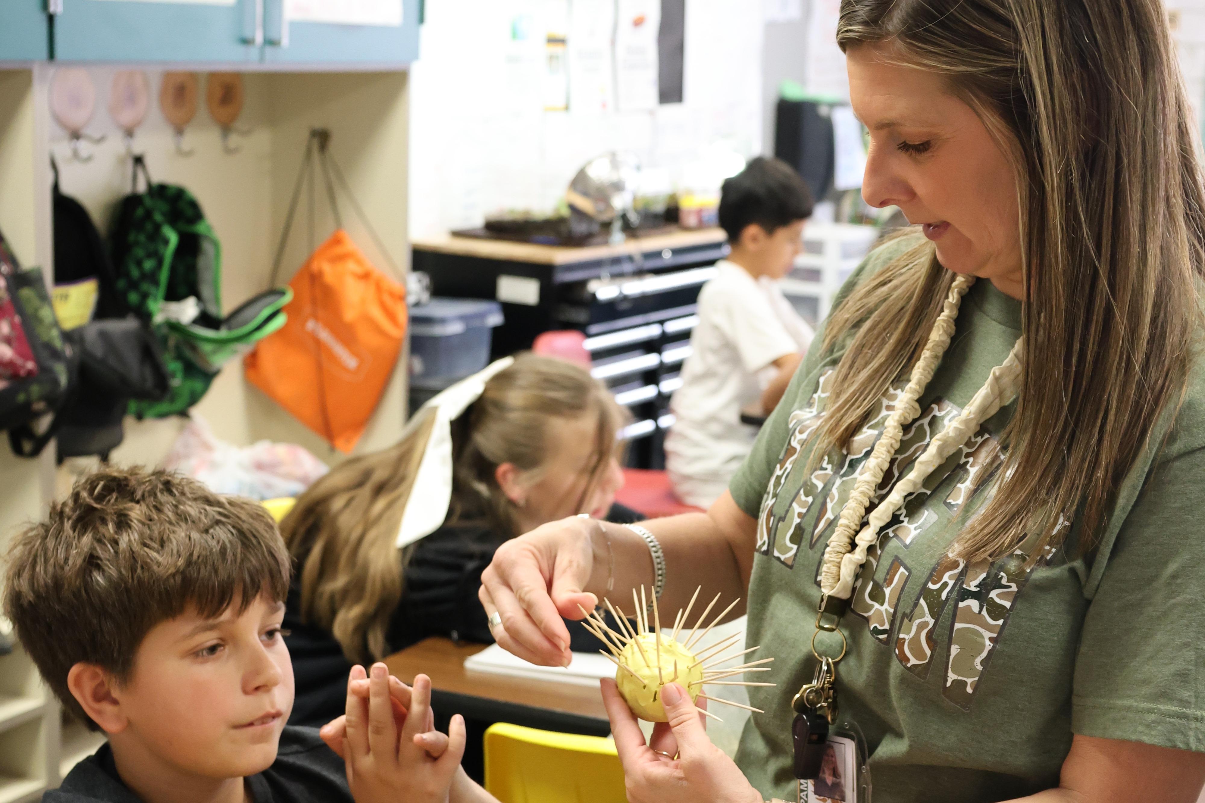 A teacher assists two students seated at a table, one holding a yellow object.