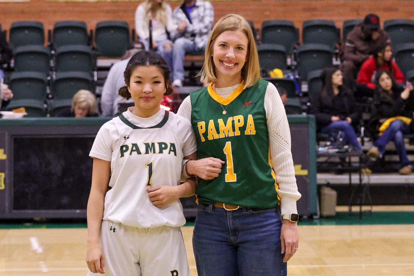 Two women stand on a basketball court, one in a white jersey and the other in green. They face the camera.