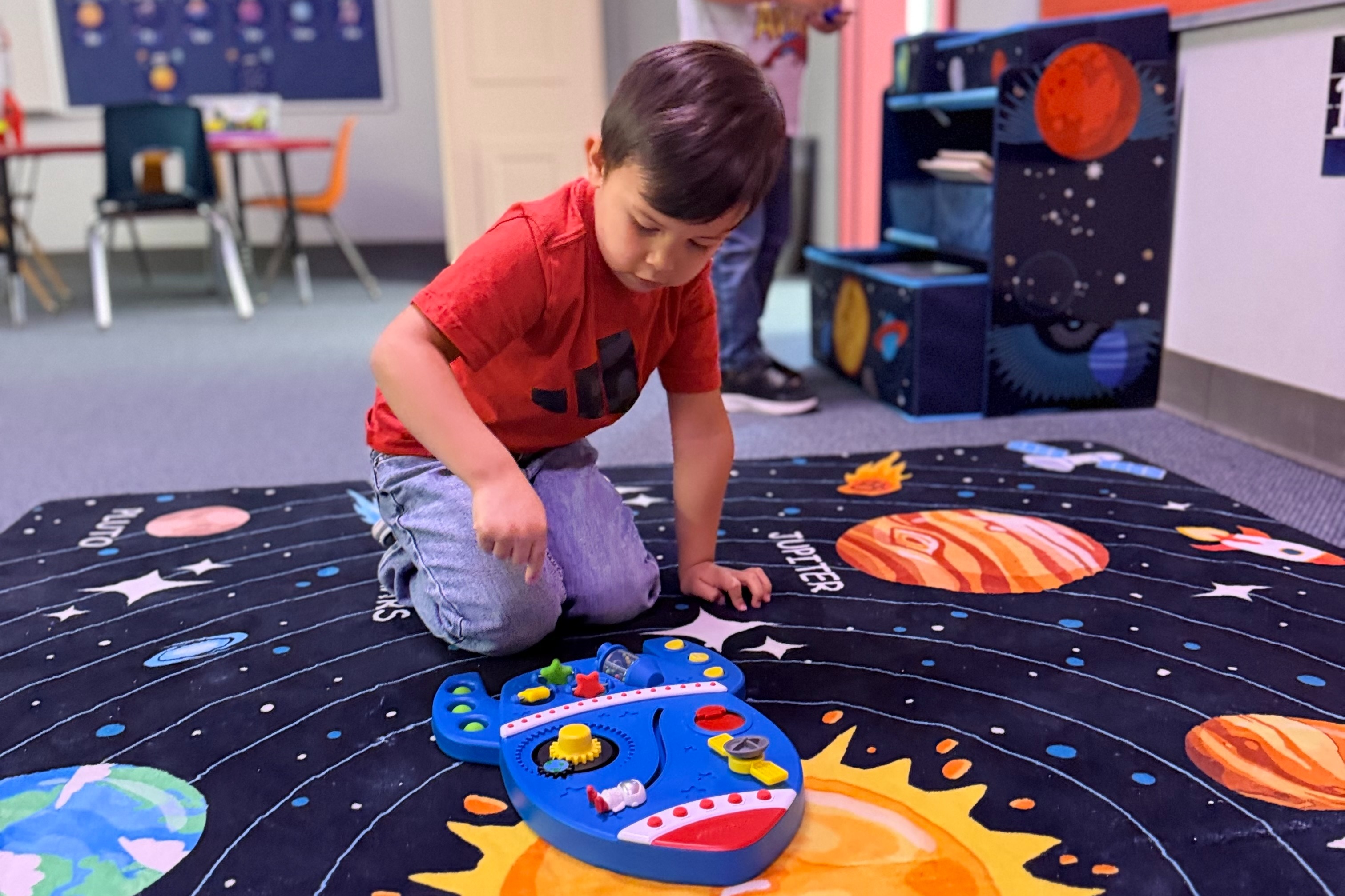 Two children play on a space-themed rug, with one kneeling to examine a toy, while another stands near a doorway.