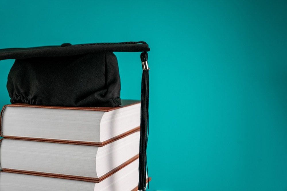 A black graduation cap sits atop a stack of white books against a teal background.