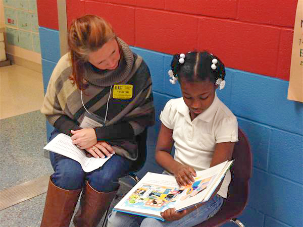 and adult volunteer reading a book in the hallway with an elementary aged student.